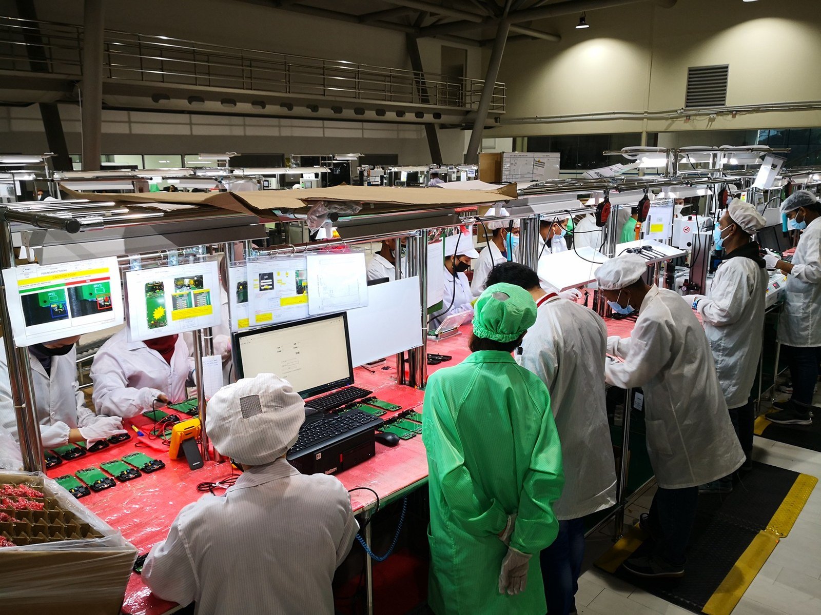A wide view of a phone circuit-board assembly hall with rows of workers in cleanroom clothing at illuminated workstations.